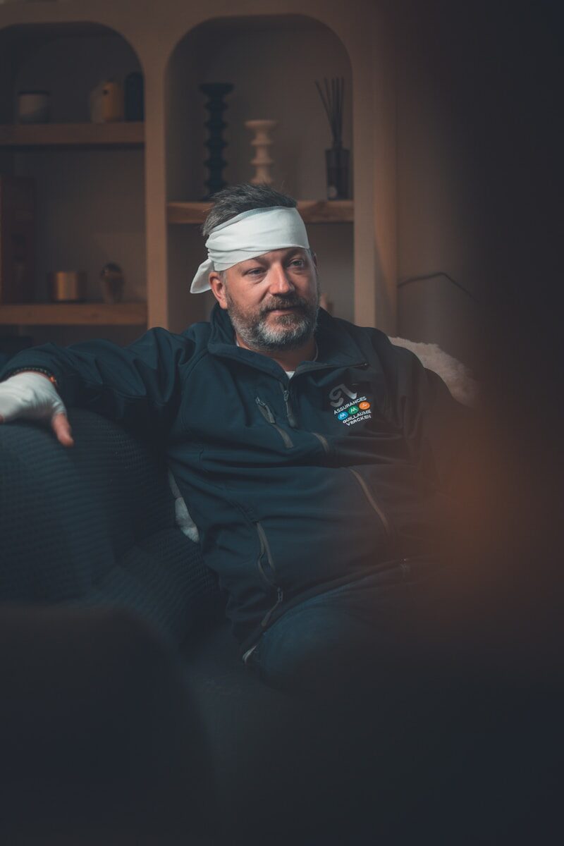 Man with bandaged head and hand sitting on couch.