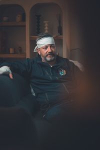 Man with bandaged head and hand sitting on couch.